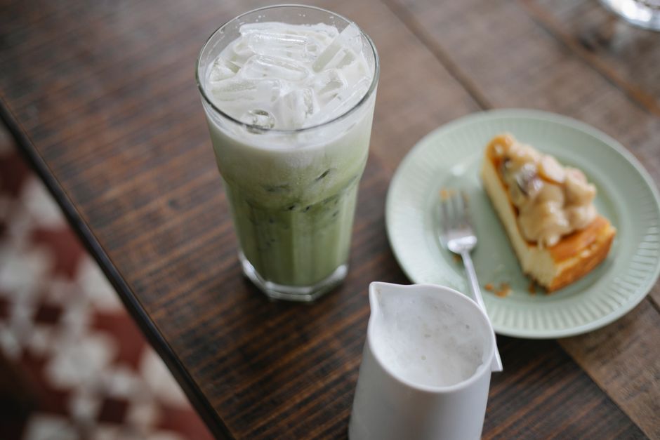 High angle tasty iced matcha tea latte placed on wooden table with yummy sweet cake and milk jigger