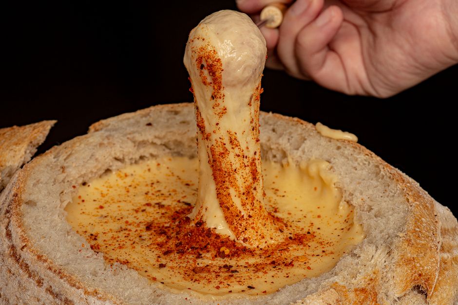 Delicious cheese fondue in bread bowl, seasoned with spices, being dipped by a hand.