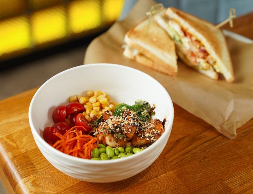 A vibrant salad bowl with grilled chicken, vegetables, and a side of sandwiches on a wooden table.